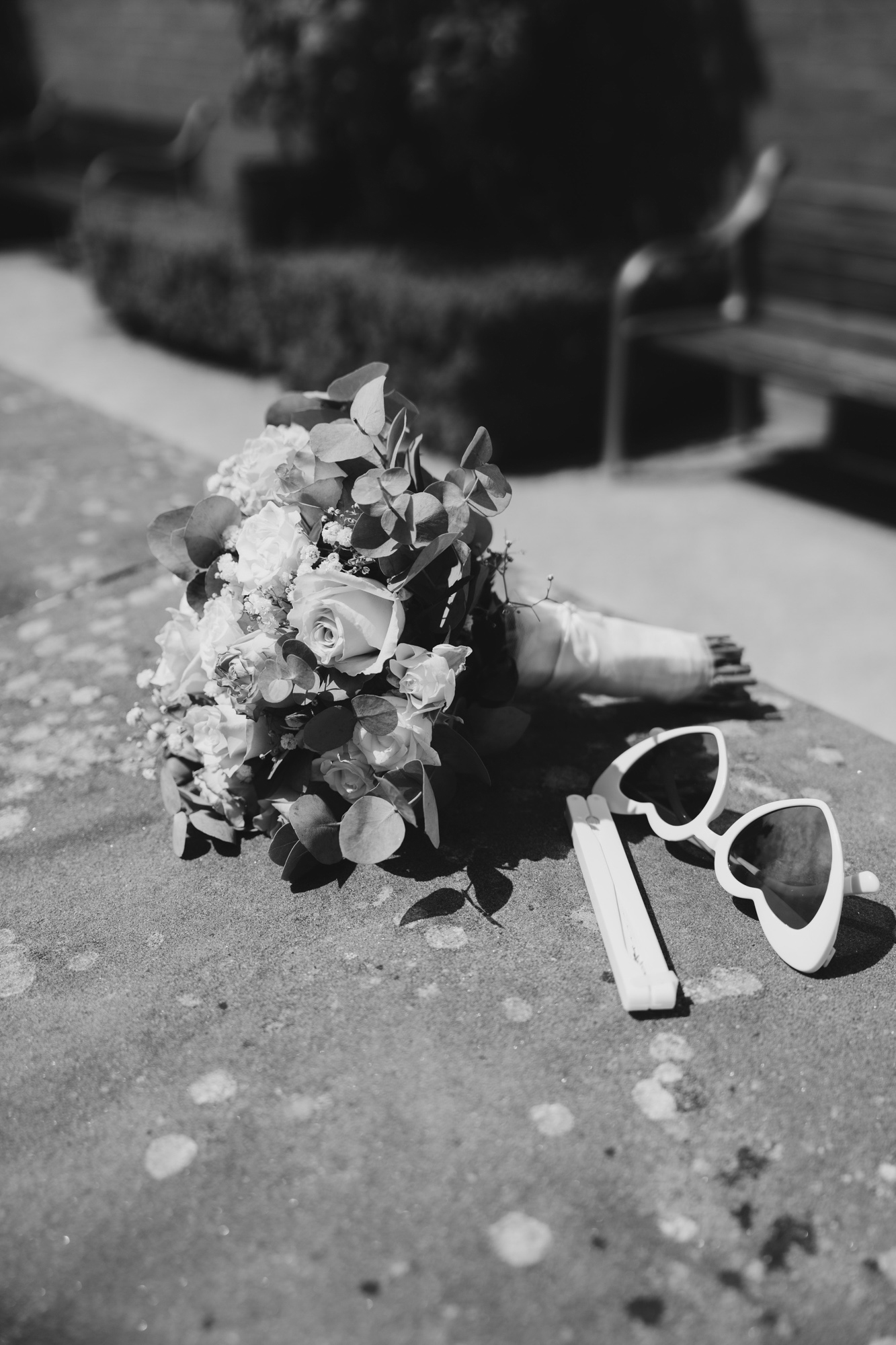Black and white photo of a bridal bouquet and heart-shaped sunglasses resting on a stone surface outdoors.