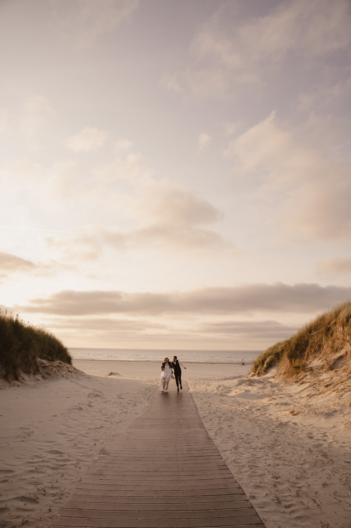 Newlywed couple walking hand in hand on a wooden path through sand dunes toward the beach at sunset.