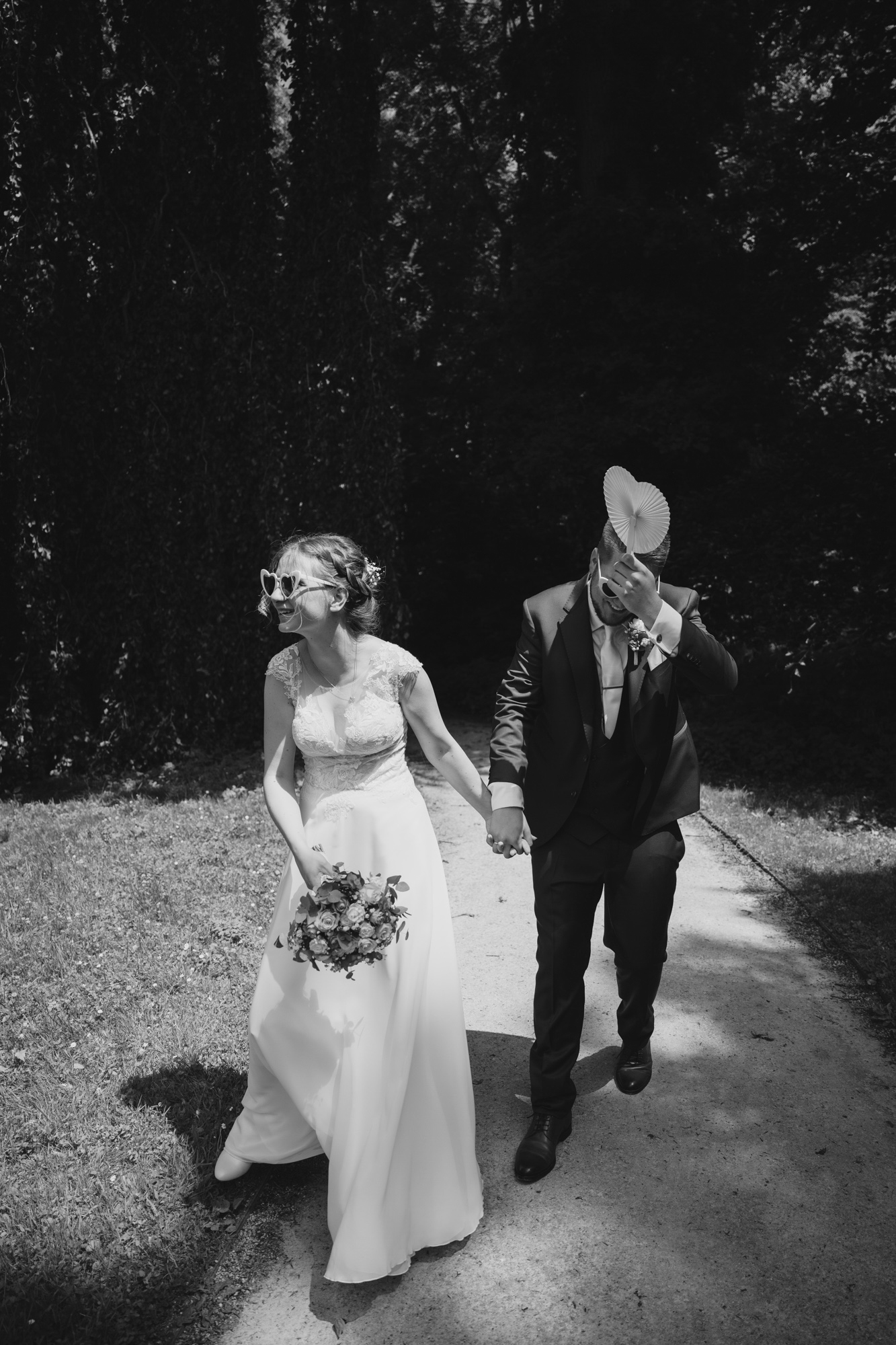 Bride and groom laughing and holding hands while walking outdoors, bride wearing heart-shaped sunglasses and holding a bouquet, groom holding a paper fan to his face.