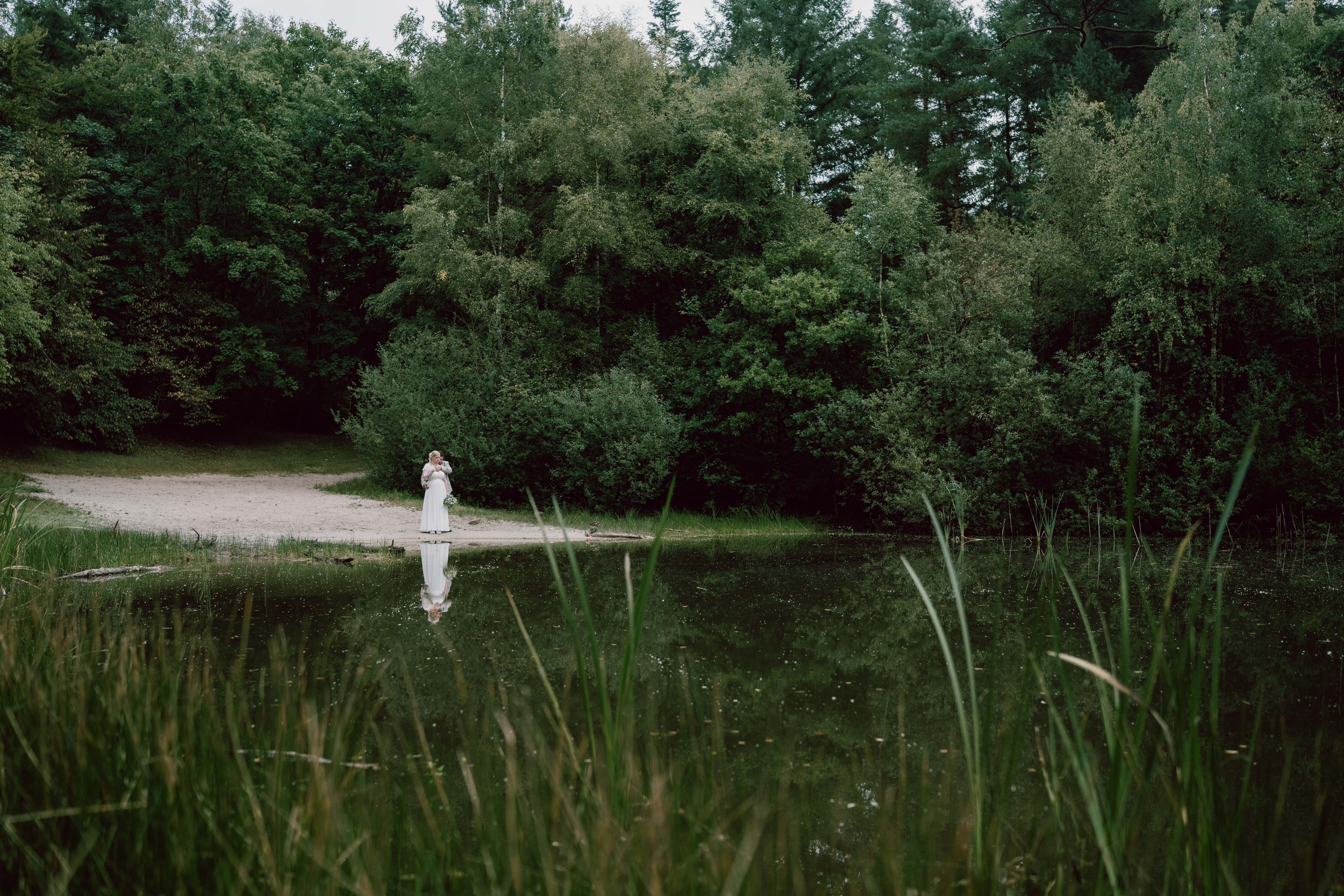 Couple in wedding attire embracing near a forest pond with their reflection visible in the water.
