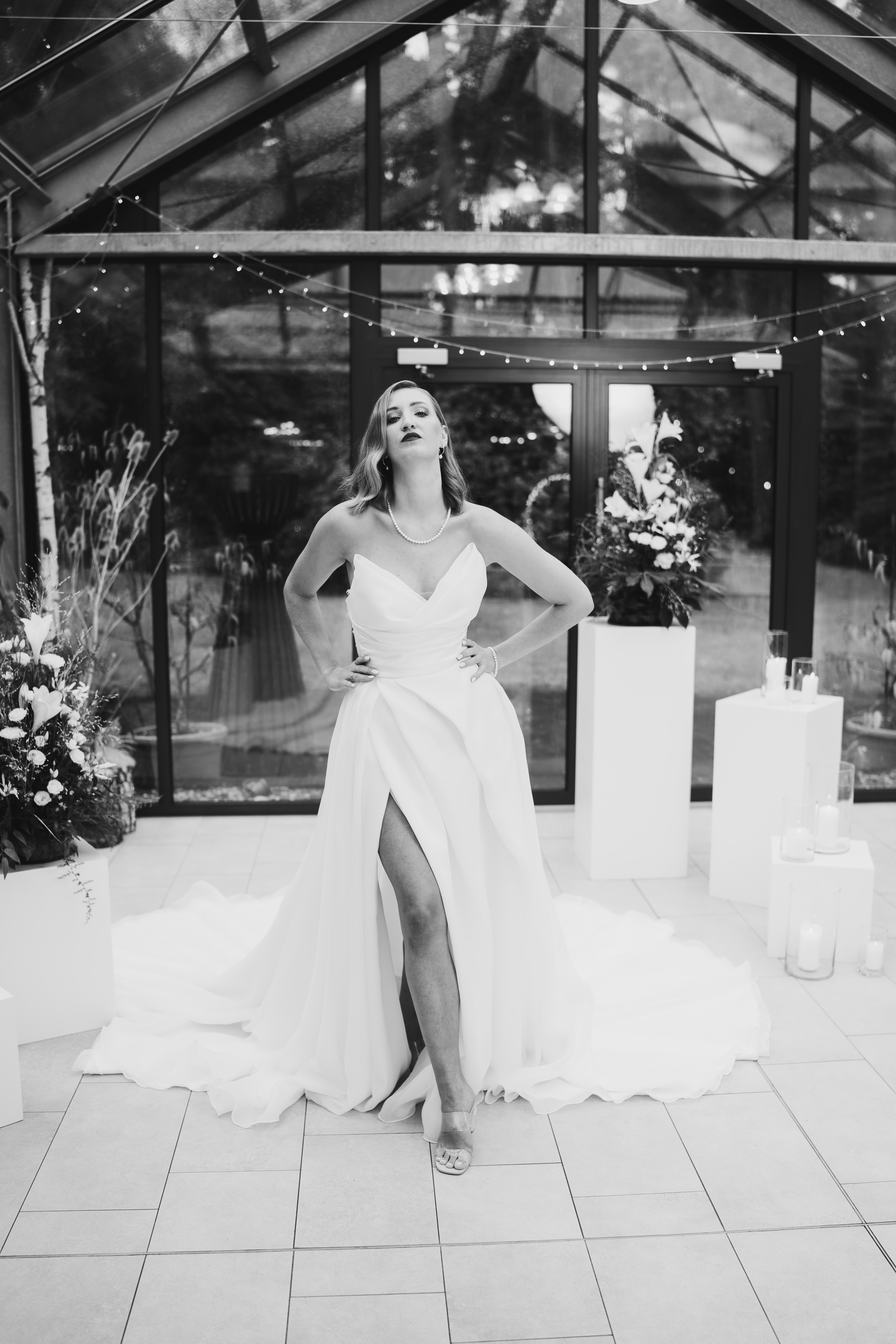 Bride in a strapless wedding dress with a thigh-high slit standing confidently in front of glass doors adorned with floral arrangements and candles.