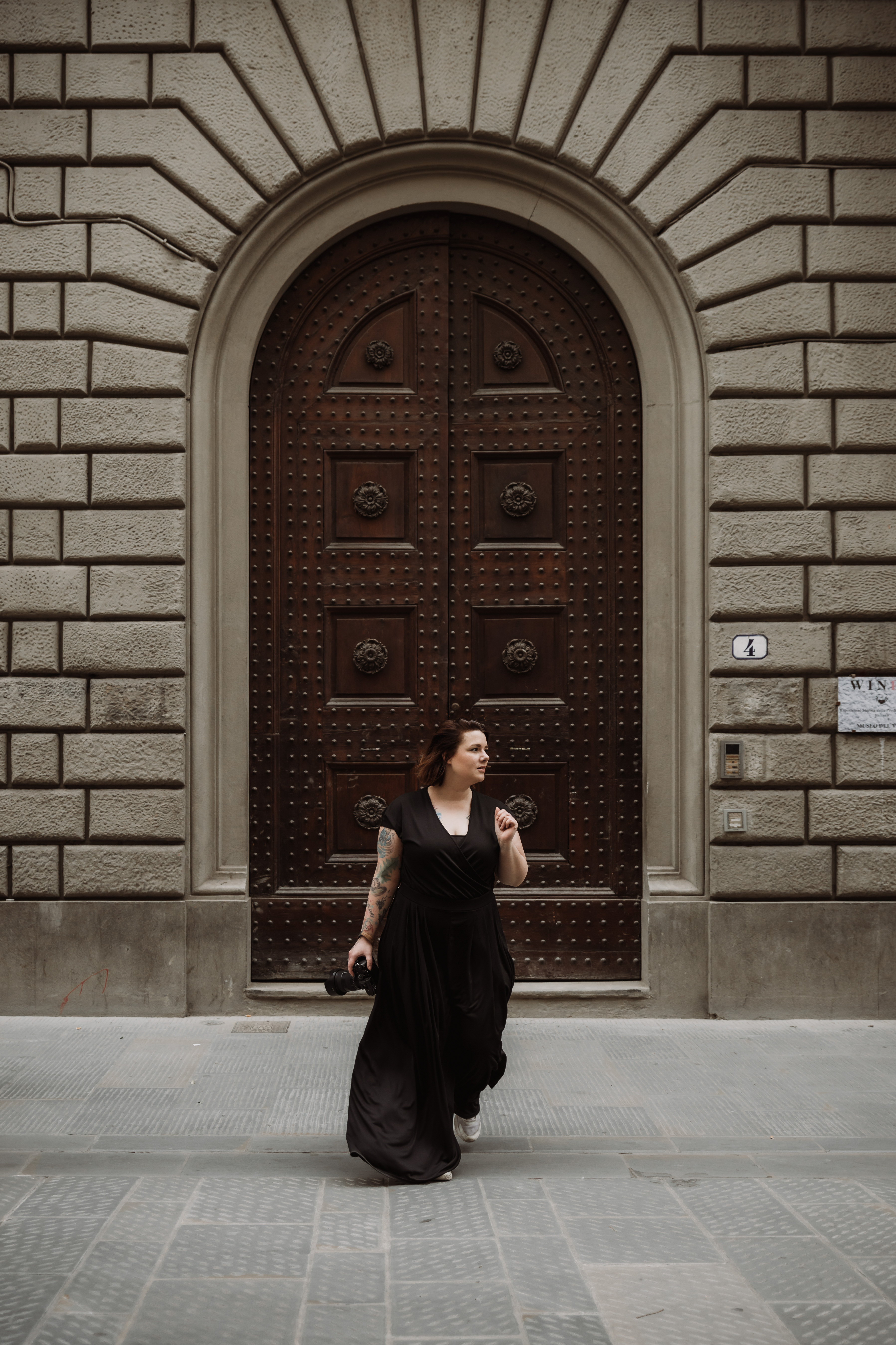 Woman in a black dress holding a camera walking in front of a large ornate wooden door set in a stone building.