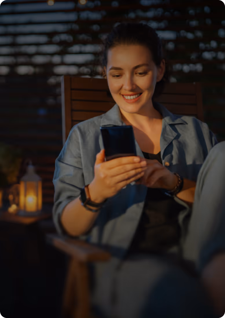 Smiling woman sitting on wooden chair and looking at her smartphone in dimly lit outdoor setting with lantern in background.