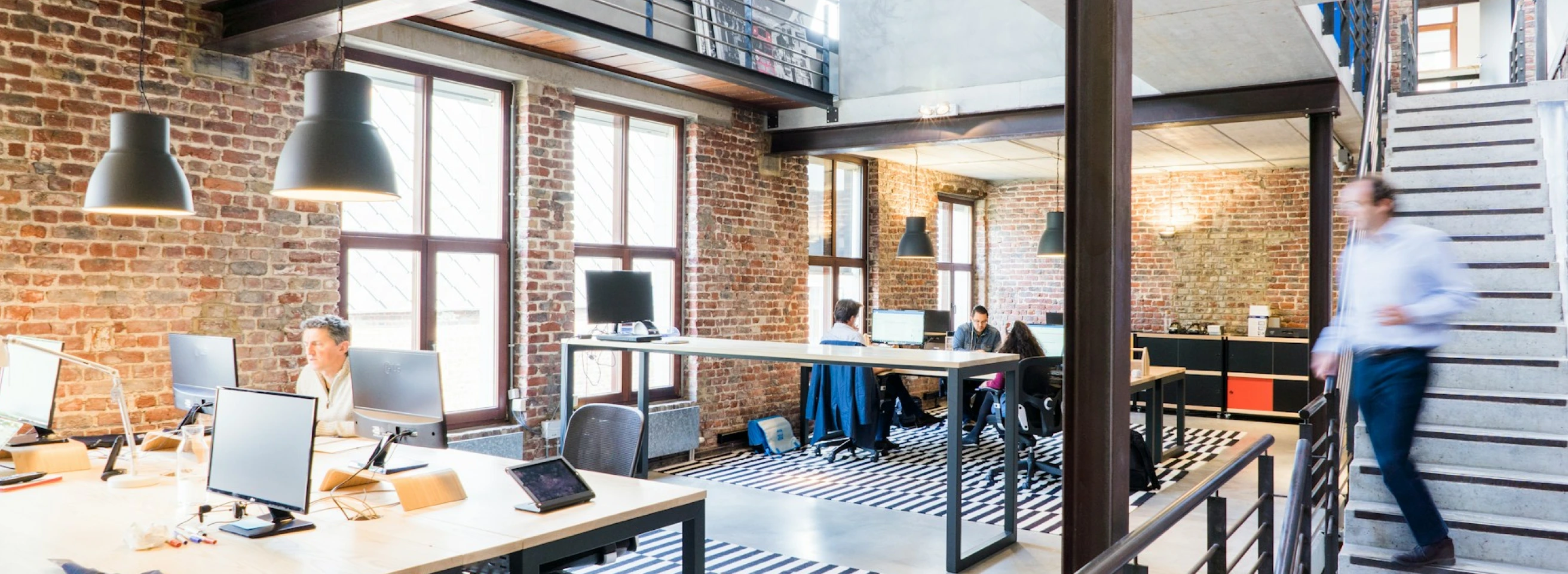 Mitarbeitende arbeiten an Schreibtischen in einem modernen Büro mit Industrie-Loft-Charakter, im Hintergrund eine Person auf der Treppe