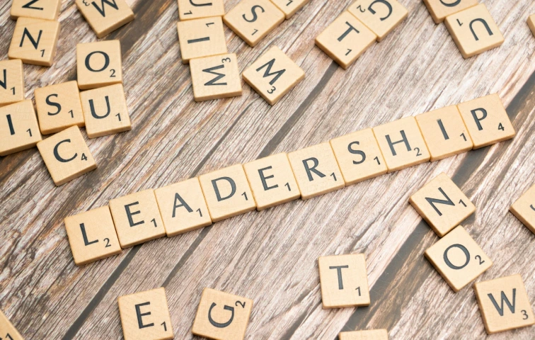 Scrabble-Buchstabensteine bilden auf verwittertem Holzuntergrund das Wort 'Leadership', umgeben von losen Einzelbuchstaben.