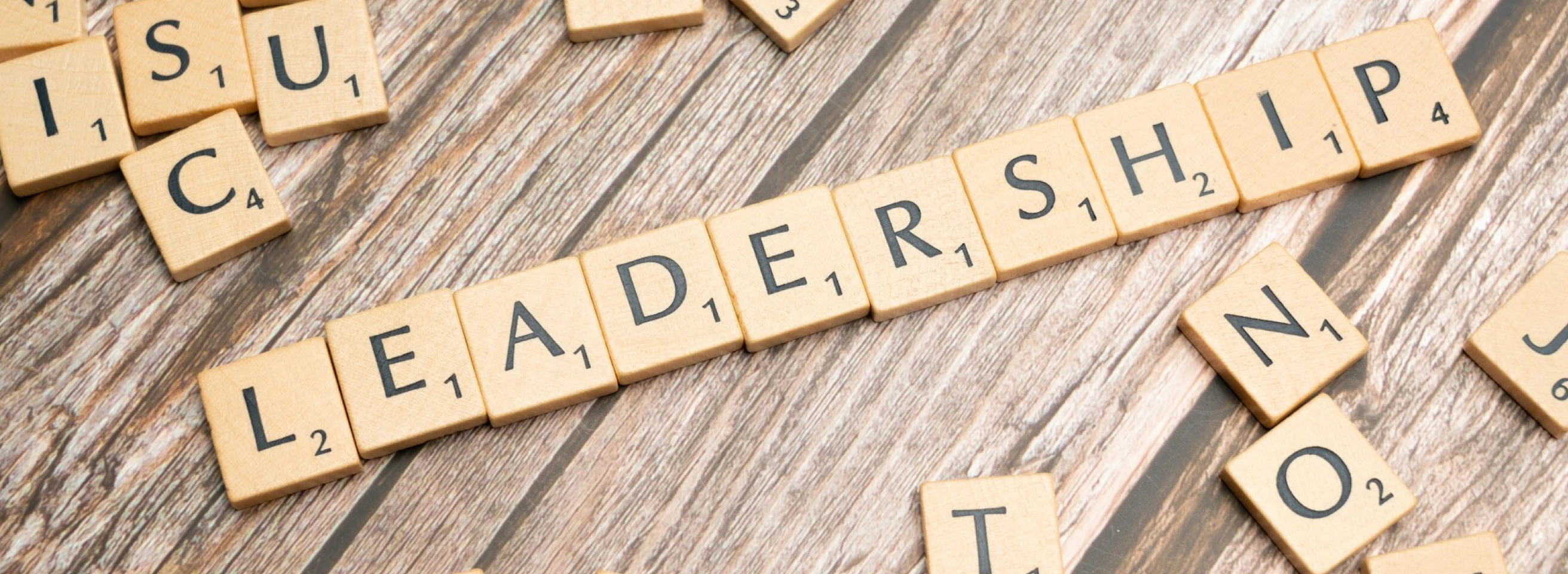 Scrabble-Buchstabensteine bilden auf verwittertem Holzuntergrund das Wort 'Leadership', umgeben von losen Einzelbuchstaben.