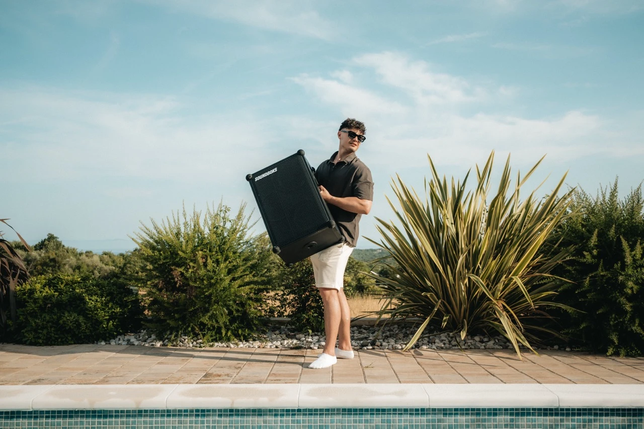 Mann mit Sonnenbrille trägt einen SOUNDBOKS Lautsprecher über die Steinterrasse am Pool.