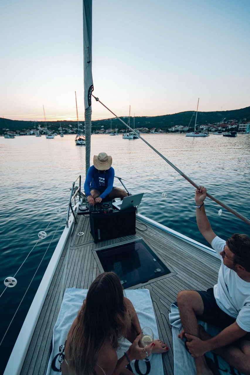 DJ mit Strohhut legt am Bug eines Segelboots auf, zwei Gäste entspannen mit Getränken an Deck.