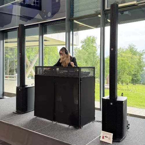 Frau mit Kopfhörern legt hinter einem schwarzen DJ-Pult zwischen zwei Säulenlautsprechern in einem verglasten Foyer auf.