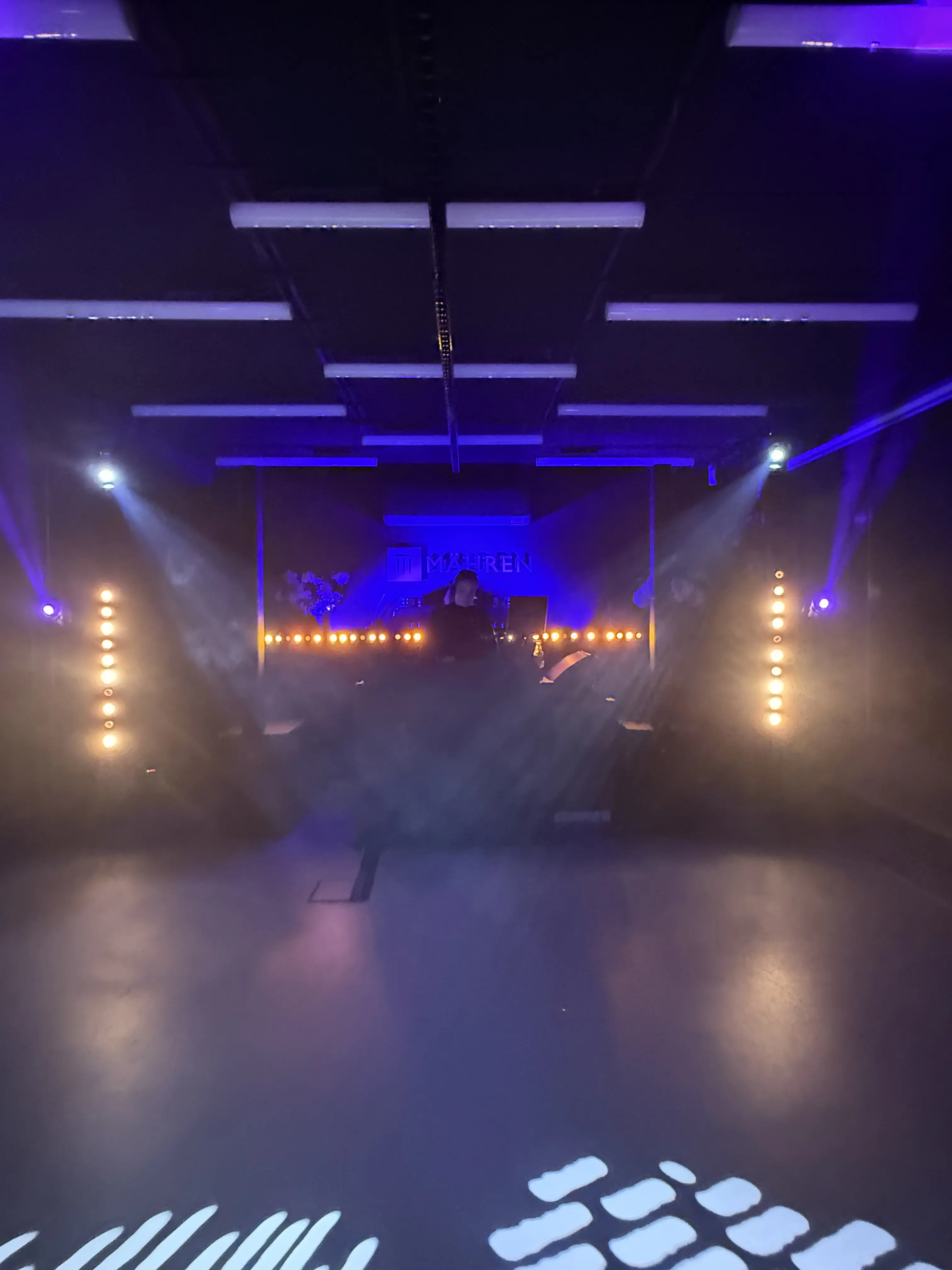 Leere Tanzfläche vor einem DJ-Pult in blauem Licht mit Nebel, Lichtstrahlen und warm leuchtenden Spots.