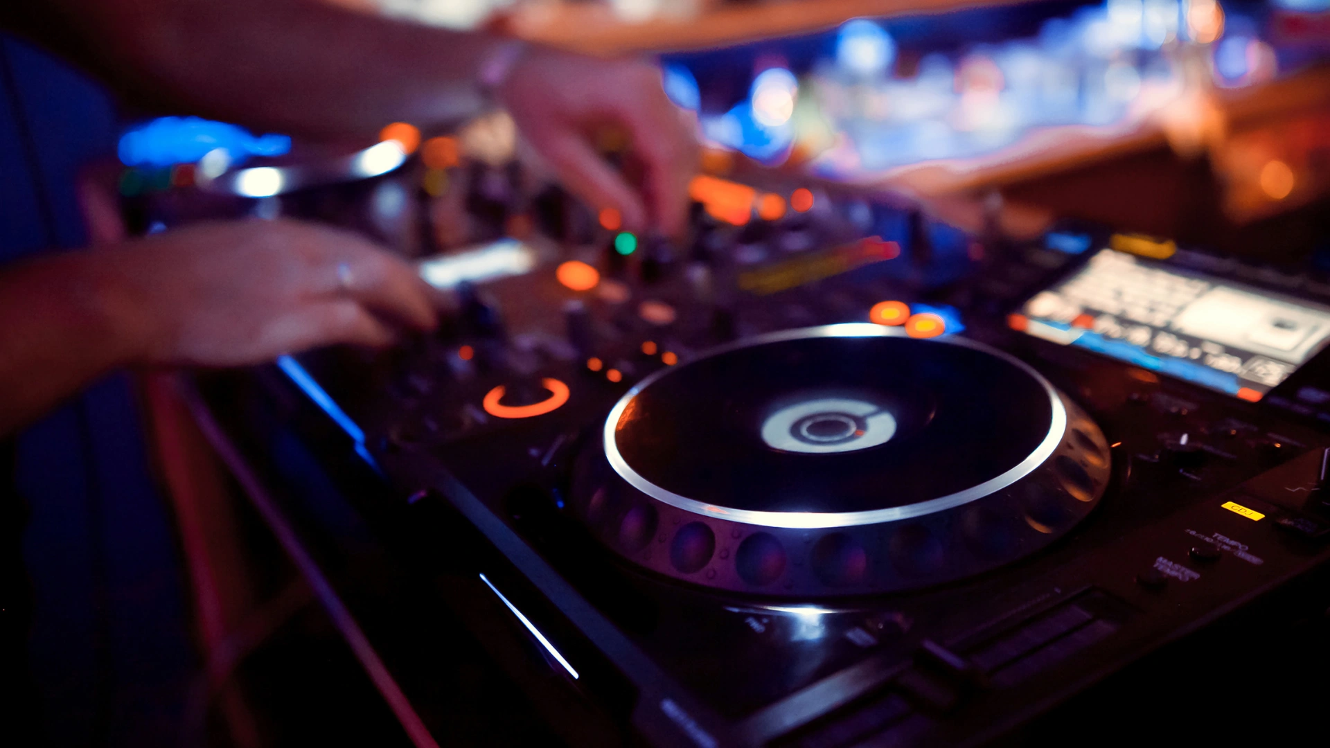 DJ legt an einem Player mit leuchtendem Jogwheel auf, im Hintergrund bunte Lichter einer Bar.