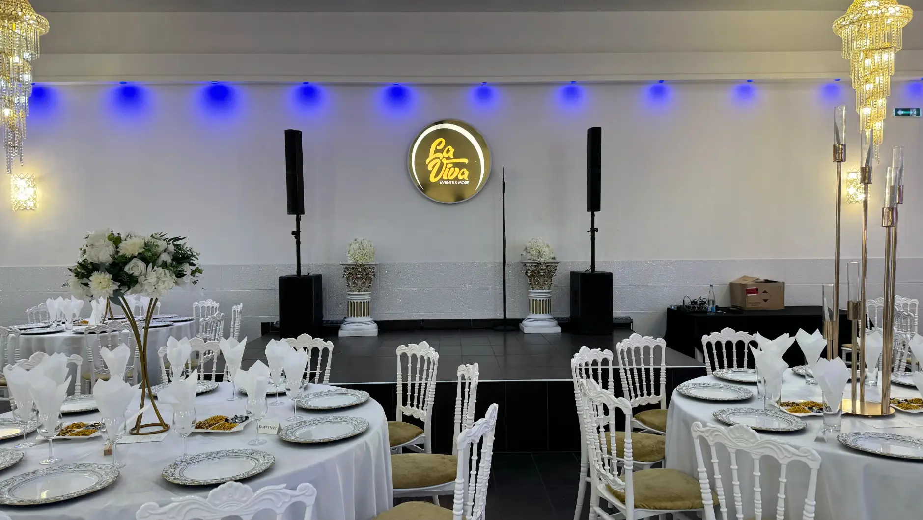 Festlich dekorierter Veranstaltungsraum mit runden Tischen, weißen Stühlen, silbernen Tellern und Bühne mit Lautsprechern.
