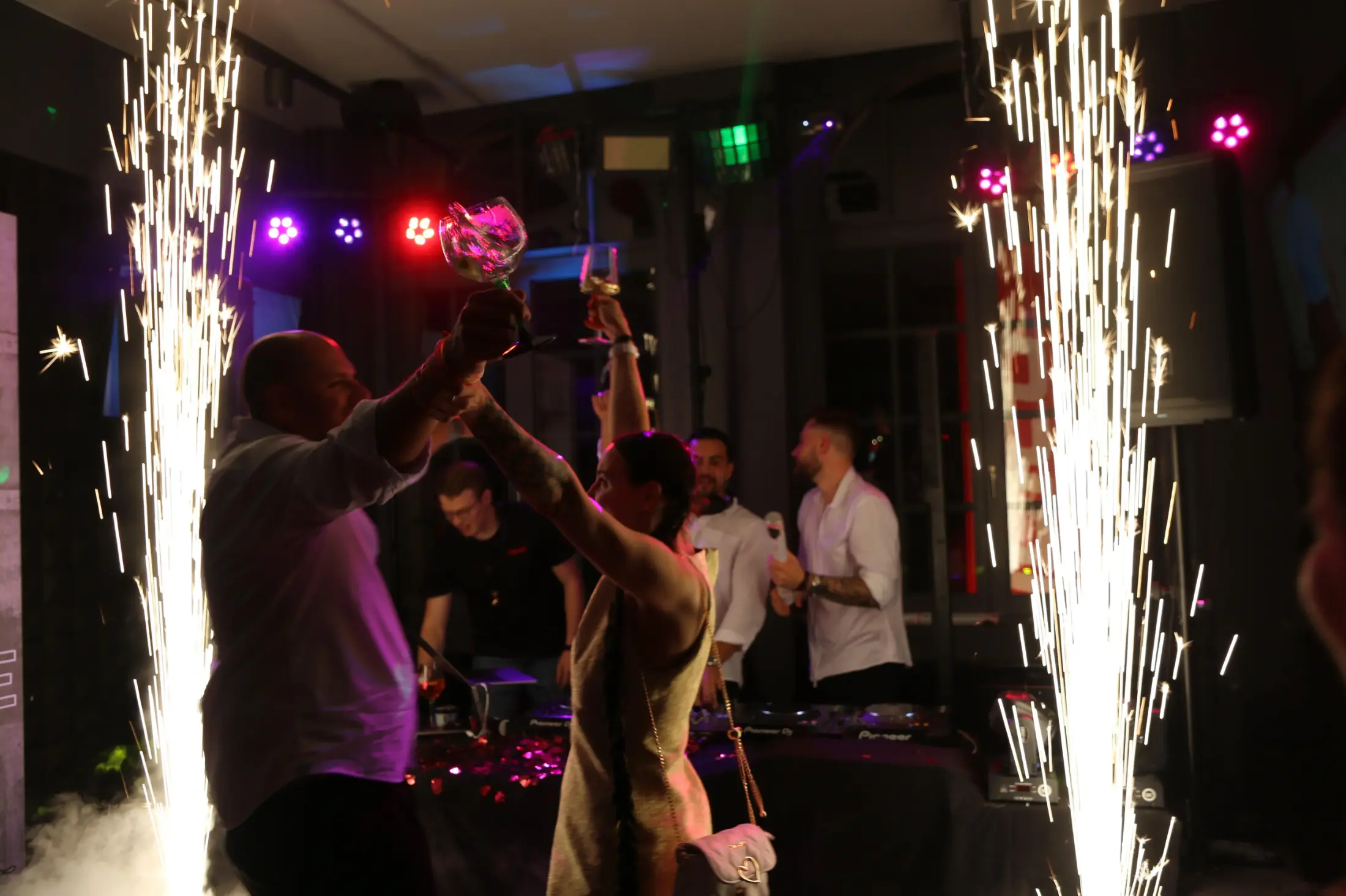 Menschen feiern mit erhobenen Gläsern vor einer DJ-Kabine, umgeben von Feuerwerksfontänen in einem Club.
