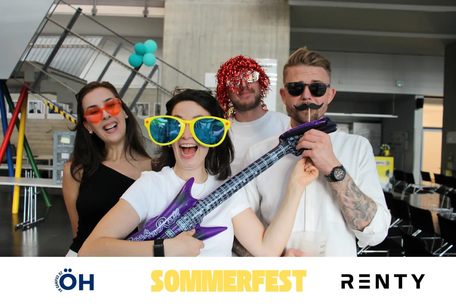 Vier ausgelassene Partygäste posieren lachend mit lustigen Fotobox-Requisiten wie einer aufblasbaren Gitarre, Perücke und bunten Riesenbrillen für ein Renty-Erinnerungsfoto auf dem Sommerfest der ÖH FH Campus 02.