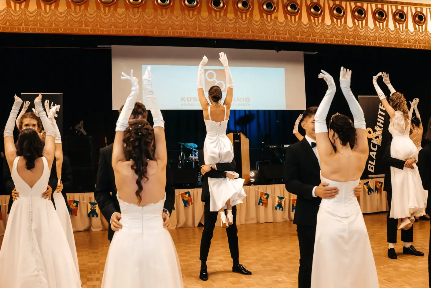 Festliche Eröffnung eines Maturaballs: Elegante Tanzpaare in weißen Abendkleidern, langen Handschuhen und dunklen Anzügen tanzen gemeinsam auf dem Parkett eines großen Festsaals.
