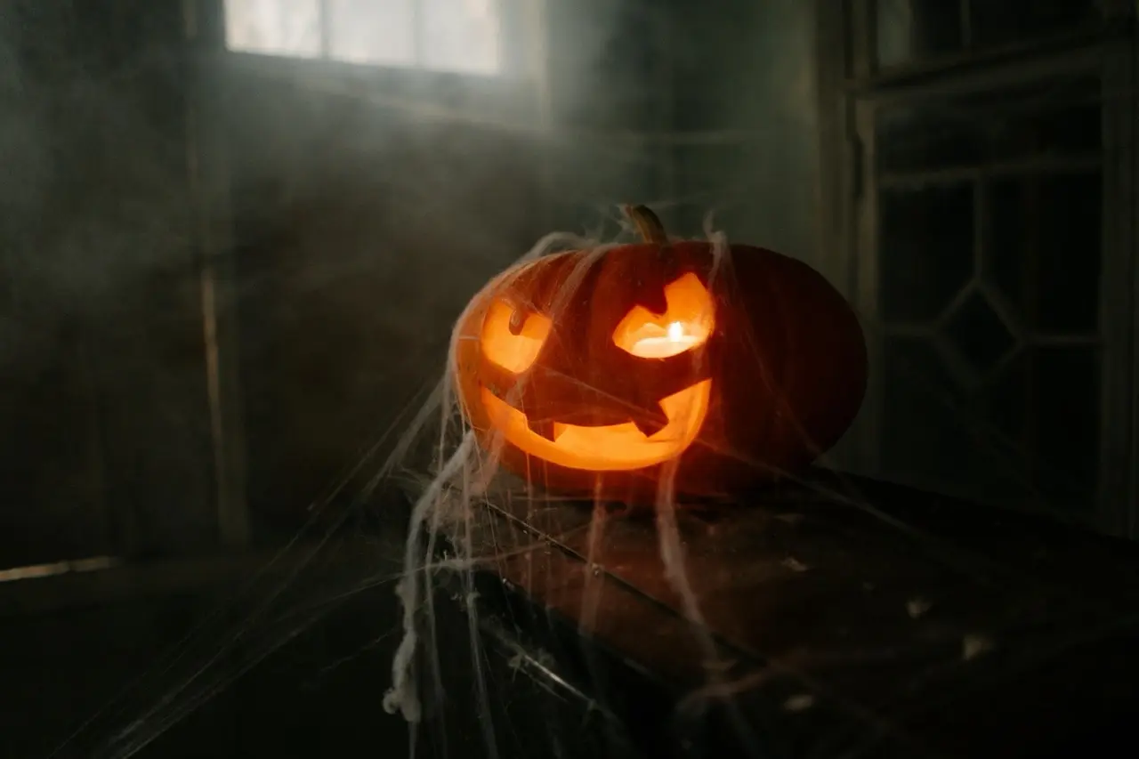 Ein klassisch geschnitzter Halloween-Kürbis mit leuchtender Fratze steht in einem dunklen, leicht vernebelten Raum, umgeben von künstlichen Spinnweben.