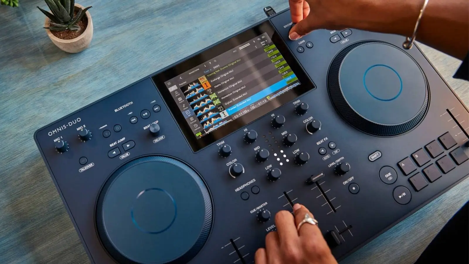 AlphaTheta OMNIS-DUO Akku-DJ-Pult in Dunkelblau mit Touchscreen, bedient auf einem blauen Tisch mit Pflanze.