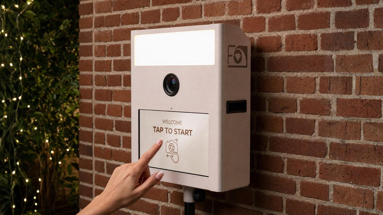 Fotobox Mini in Beige an einer Ziegelwand, deren Touchscreen mit der Hand bedient wird.