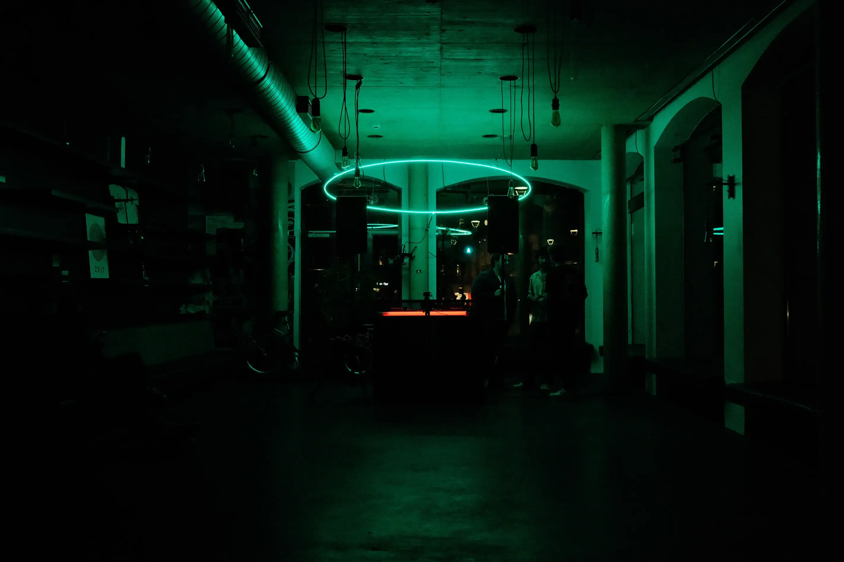 Dunkler Raum mit grünem LED-Lichtring über einem DJ-Pult am Ende des Gangs als Veranstaltungstechnik für Rave mieten.