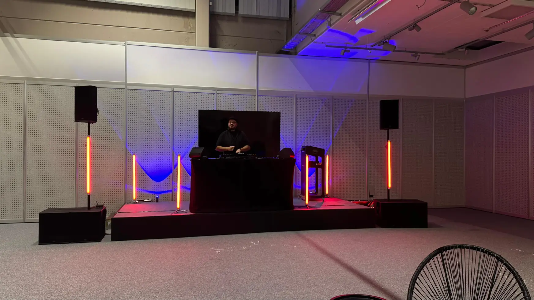 DJ am beleuchteten Pult auf einer Bühne mit zwei Boxen auf Stativen und orangen Lichtstäben als Musikanlage für Abschlussfeier mieten.