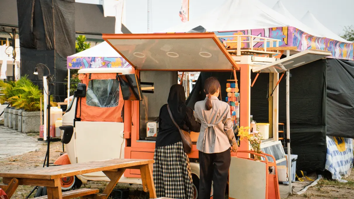 Zwei Frauen an einem orangefarbenen Marktstand im Freien neben einem Stativlicht als Veranstaltungstechnik für Messe mieten.