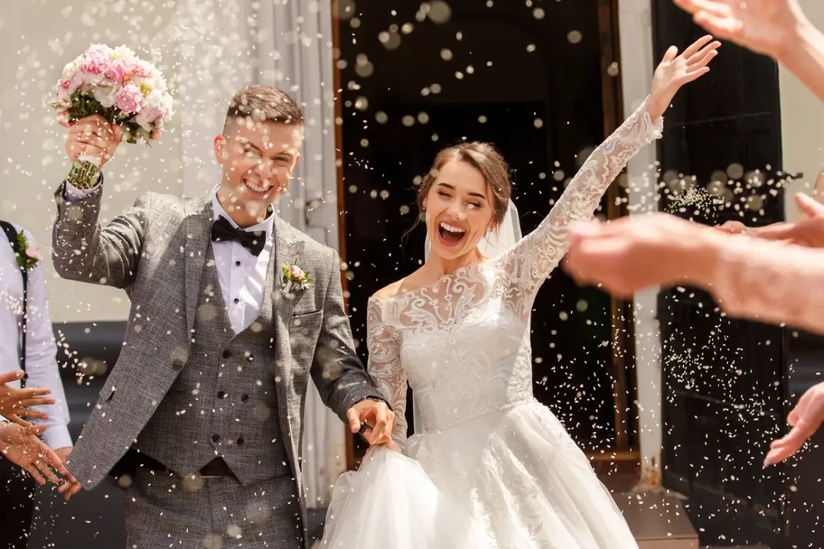 Lachendes Brautpaar wird von Gästen mit Reis beworfen vor einem Gebäude als Veranstaltungstechnik für Hochzeit mieten.