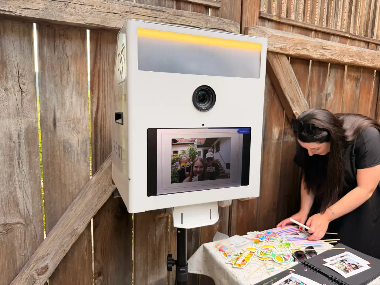 Weiße Fotobox auf einem Stativ vor einer Holzwand, daneben bereitet eine Frau an einem kleinen Tisch bunte Foto-Requisiten und ein Fotoalbum vor.