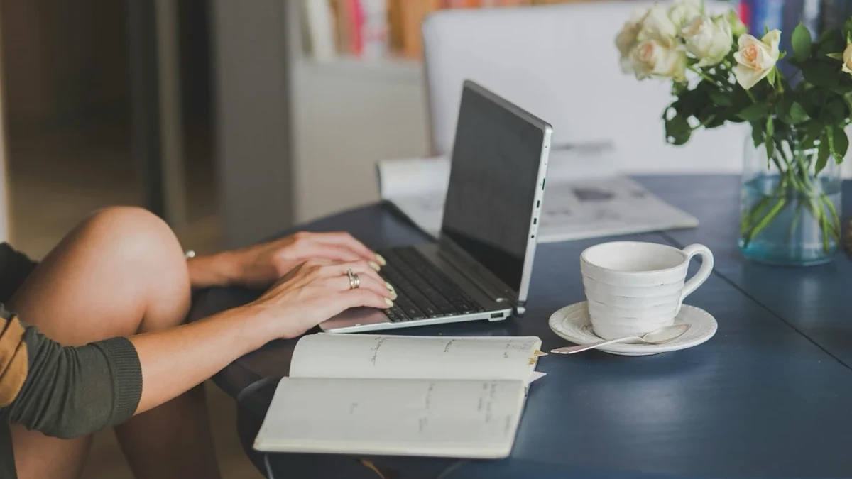 Frau formuliert am Laptop eine Einladung zum Geburtstag, daneben Notizbuch und Blumen
