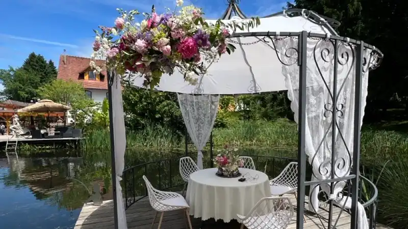 Weißer Pavillon mit üppigem Blumenschmuck und gedecktem Tisch auf einem Holzsteg am Teich