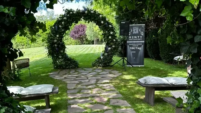 Steinweg unter einem Efeubogen führt zu einem runden Blumenbogen mit Bänken im Garten