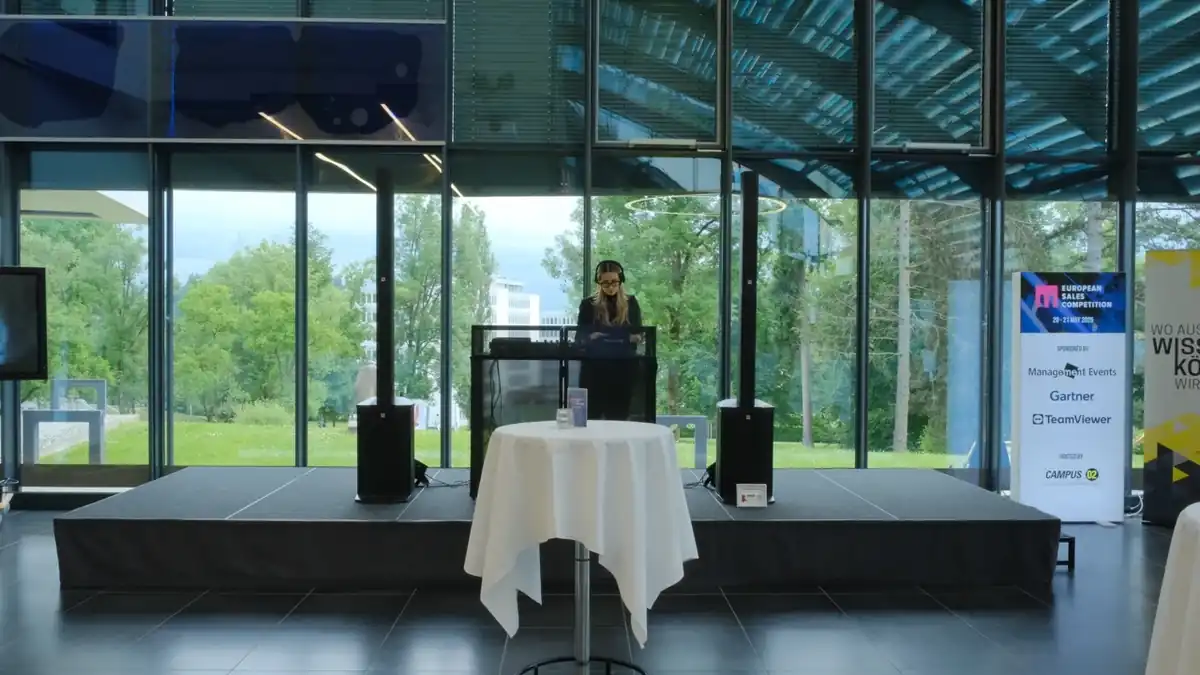 DJ auf einer Bühne in einem lichtdurchfluteten Foyer, davor Stehtische und Gäste