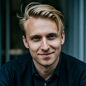 Portrait of a smiling young man with blond hair wearing a dark shirt against a blurred background.
