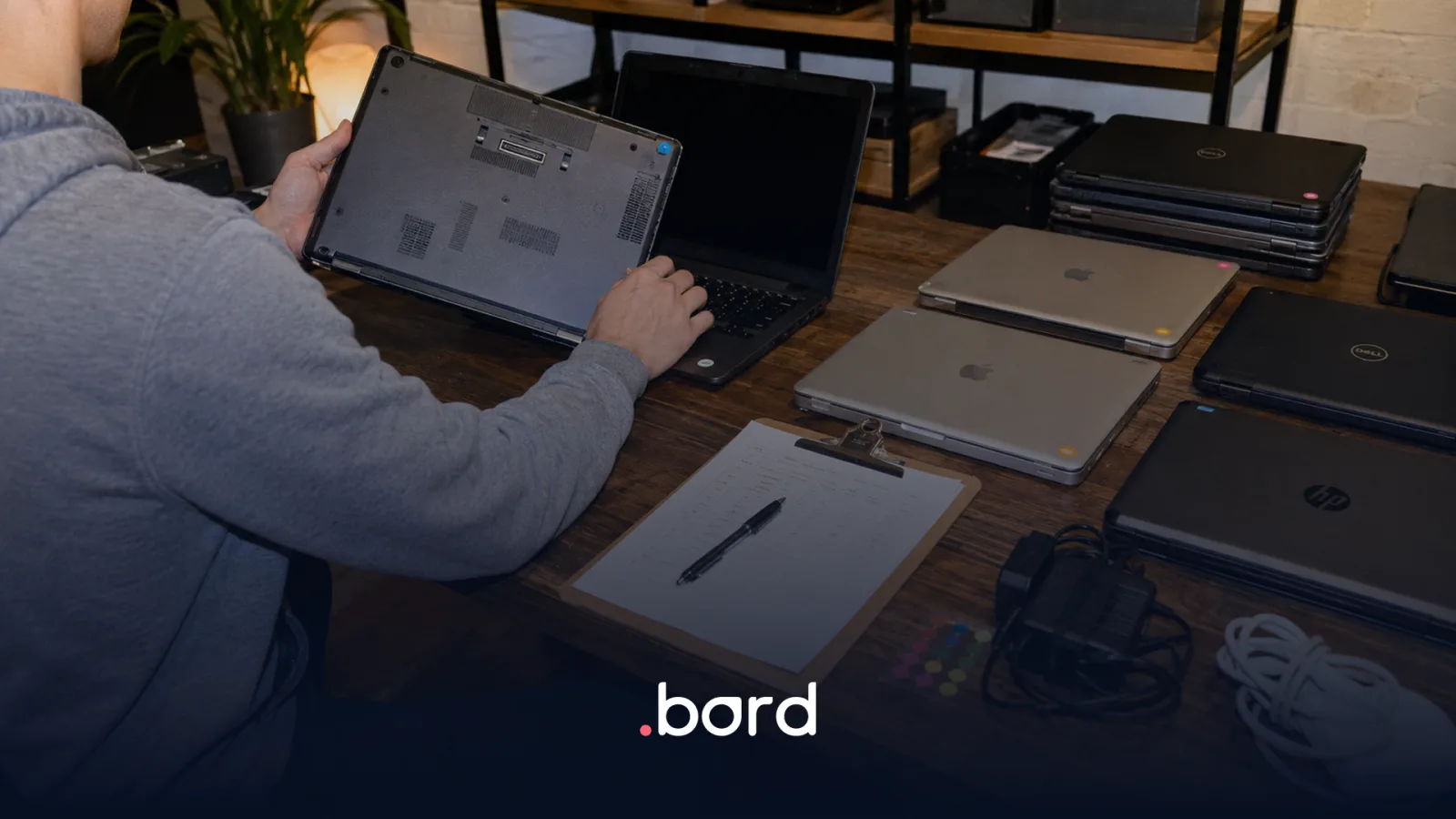 Person inspecting MacBook, Dell and HP laptops on a table with a clipboard checklist and chargers. .bord logo