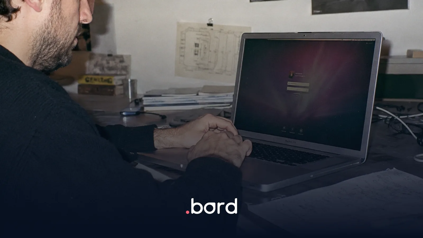 Man at a MacBook Pro showing the login screen, at his desk. .bord logo