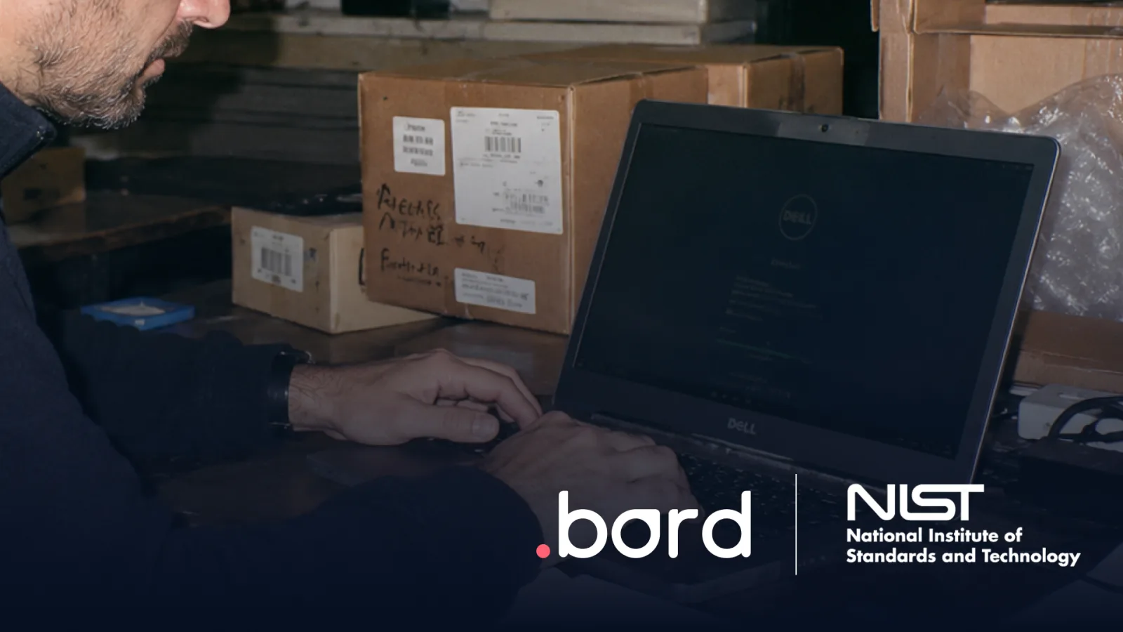 Person working on a Dell laptop among labeled boxes in a warehouse. .bord and NIST logos