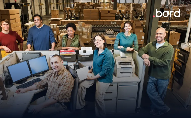Team of seven posing in an office area inside a warehouse full of boxes. .bord logo