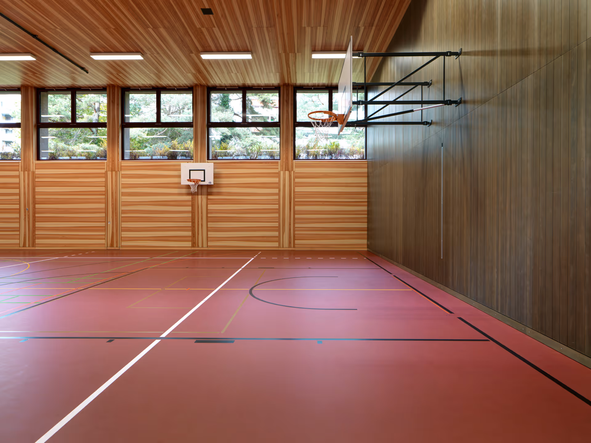 Indoor basketball court with wooden walls and ceiling, large windows, and two basketball hoops.