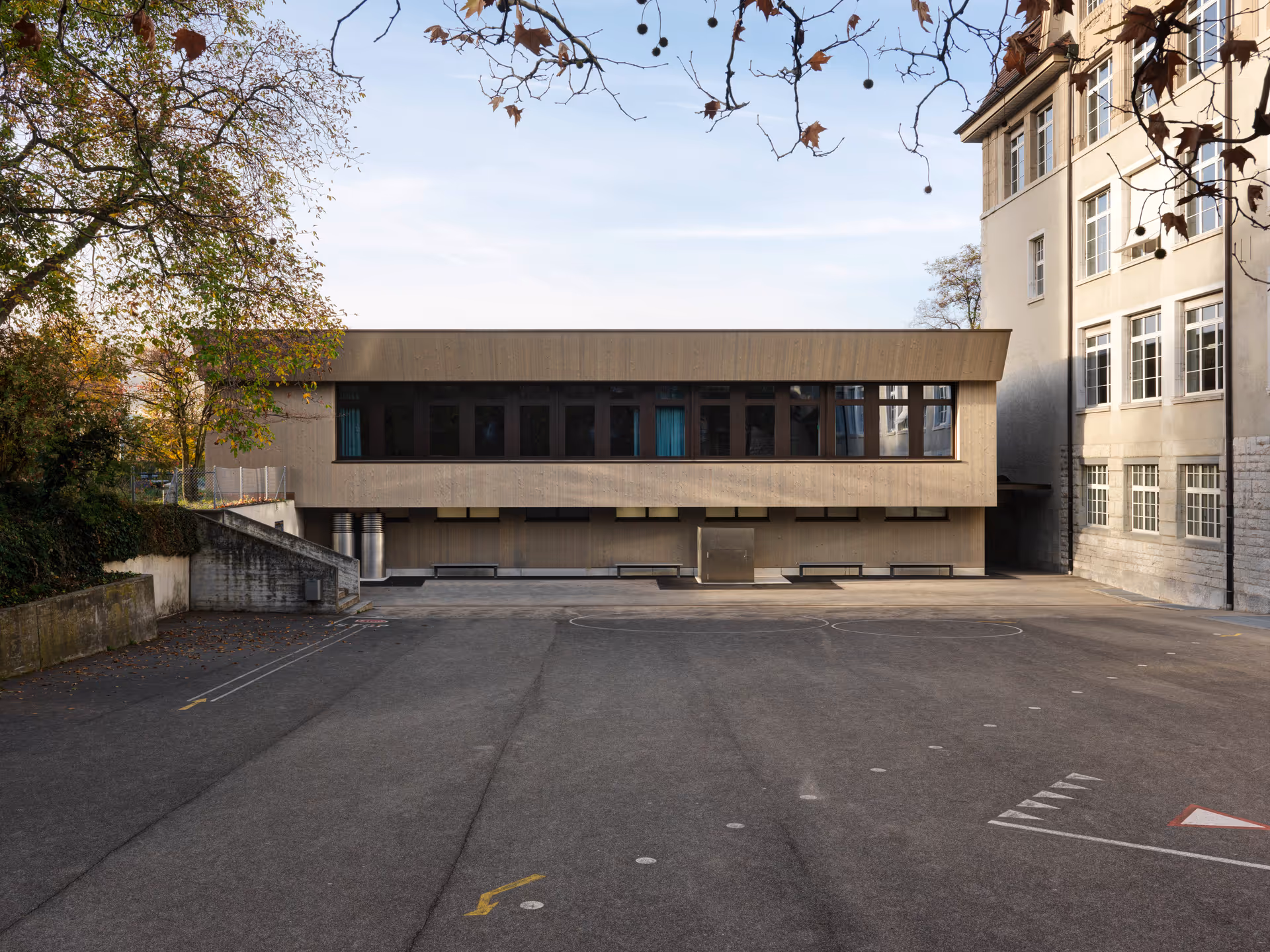 Courtyard with asphalt ground and two adjacent buildings, one modern with large dark windows and one older with many white-framed windows, under bare tree branches.
