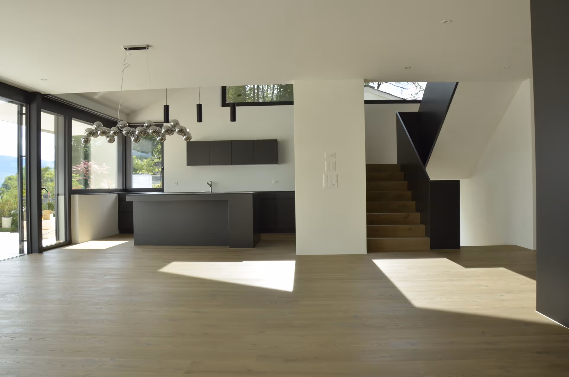 Modern open kitchen with black cabinetry and island, large windows, wooden floors, and stairs with black railing.