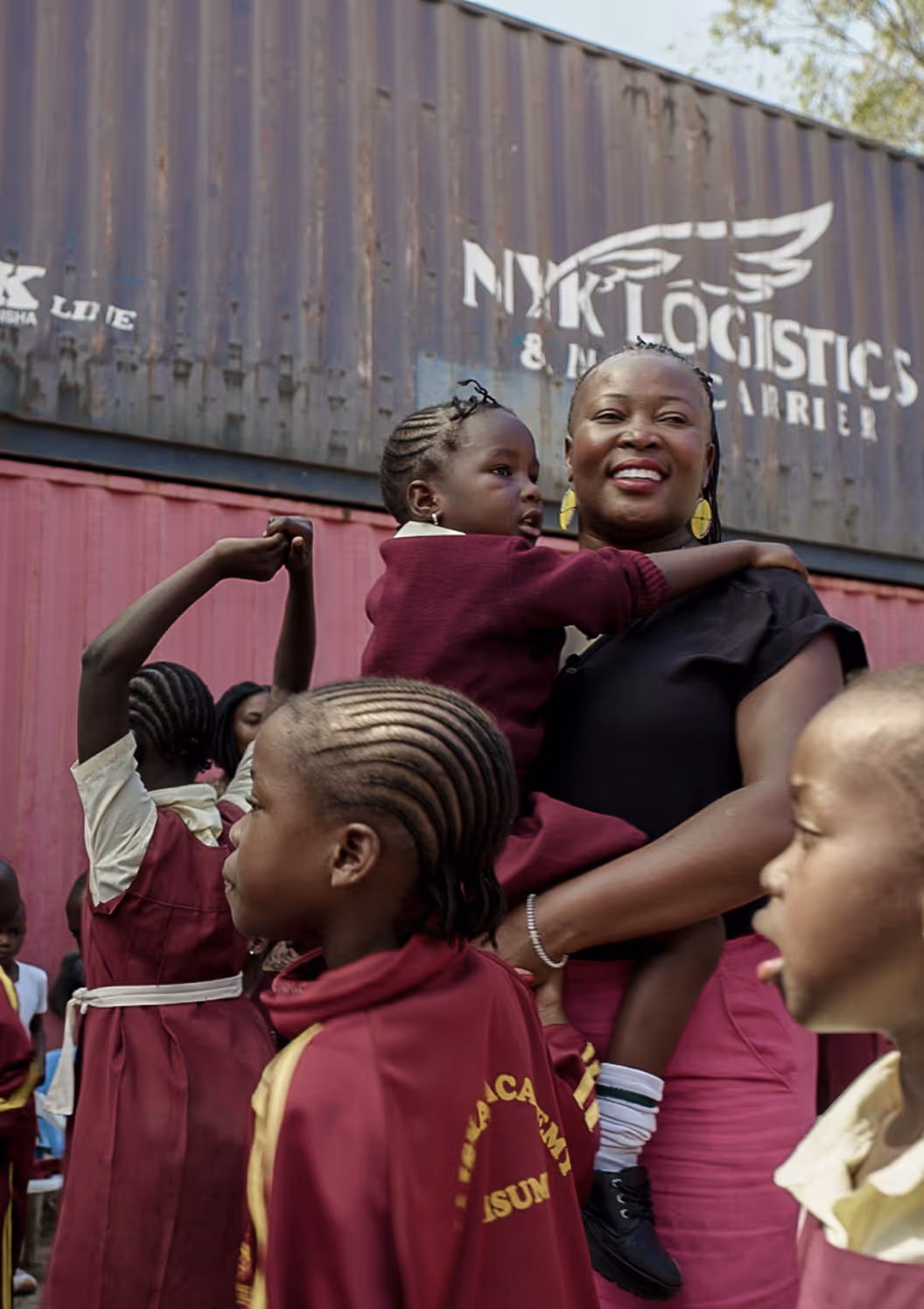 Smiling woman holding a child surrounded by children in maroon and yellow school uniforms outdoors.