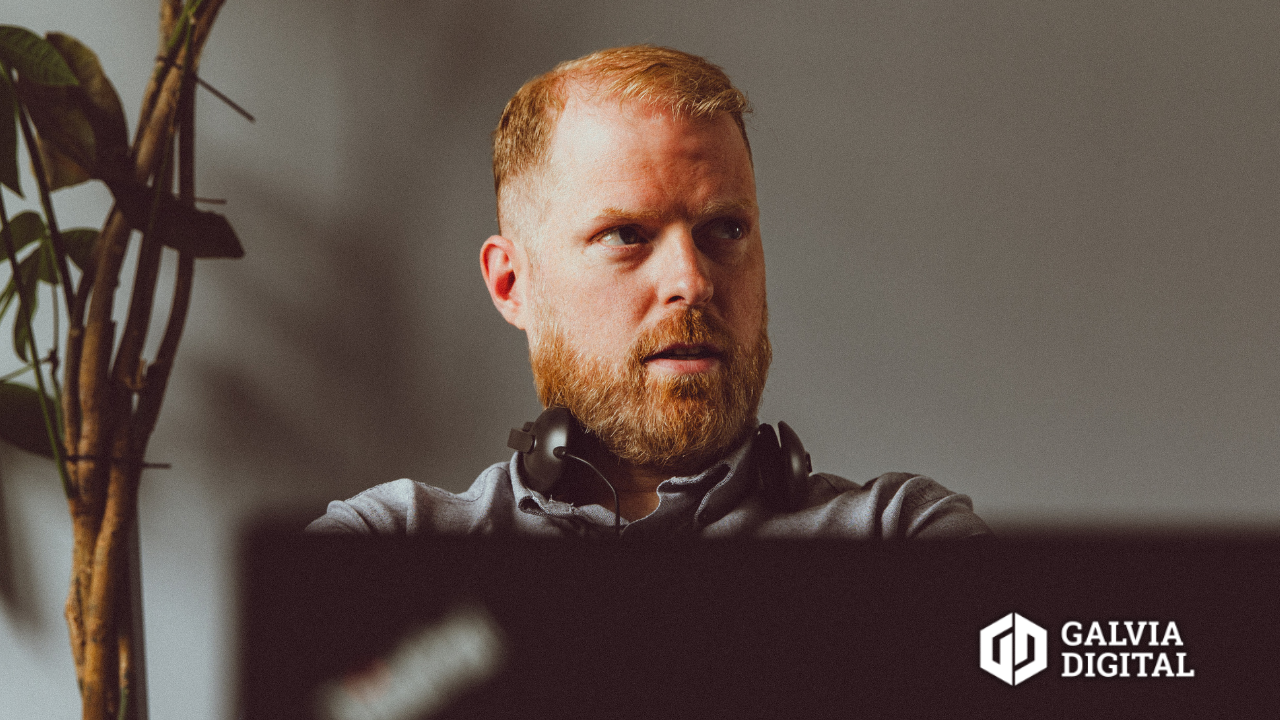 Bearded man with headphones around his neck working on a laptop with a plant in the background.