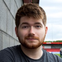 Young man with short brown hair and beard leaning against a stone wall outdoors with a stadium in the background.