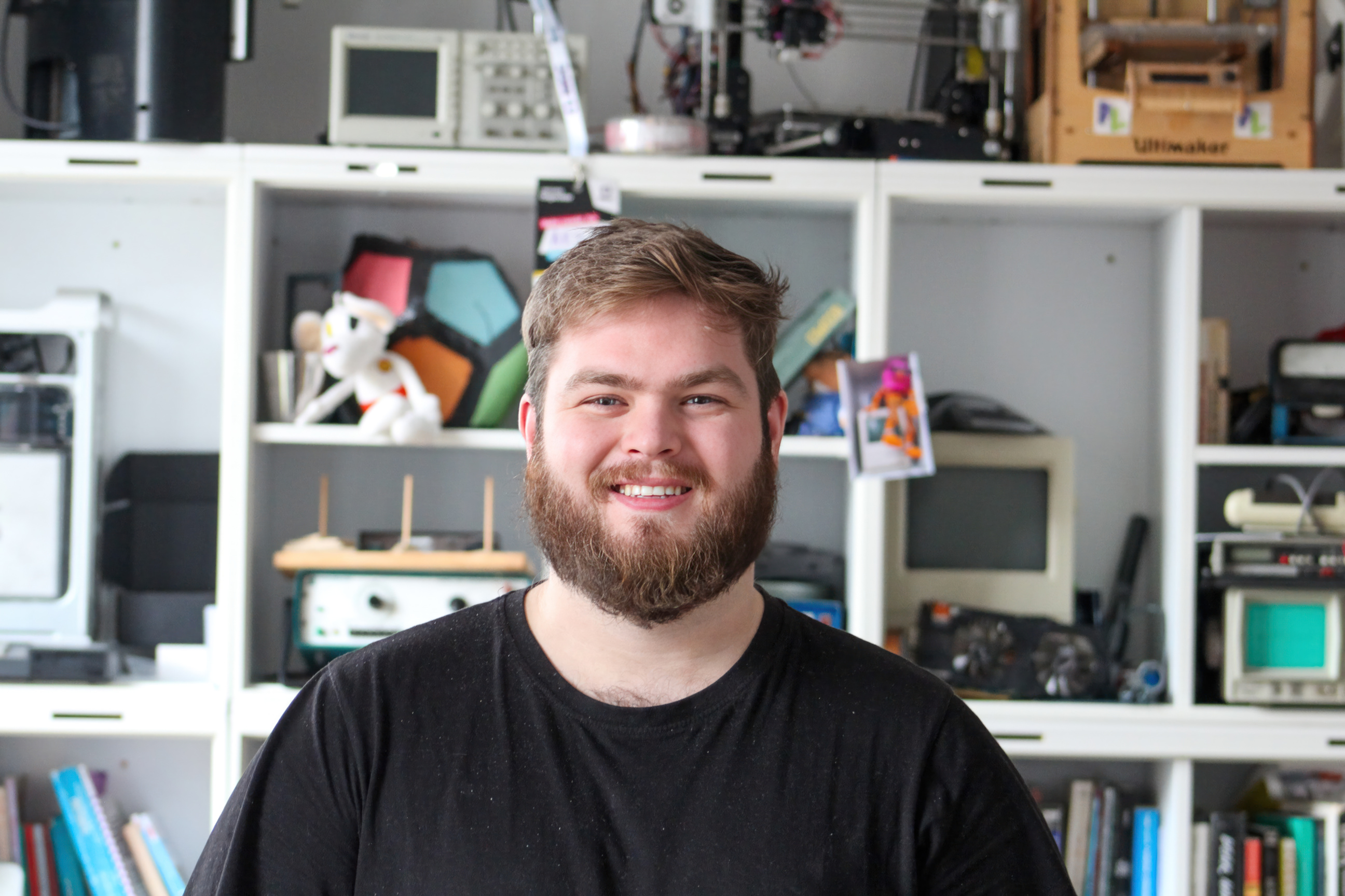 Smiling man with a beard wearing a black shirt in front of shelves filled with books, vintage electronics, and toys.
