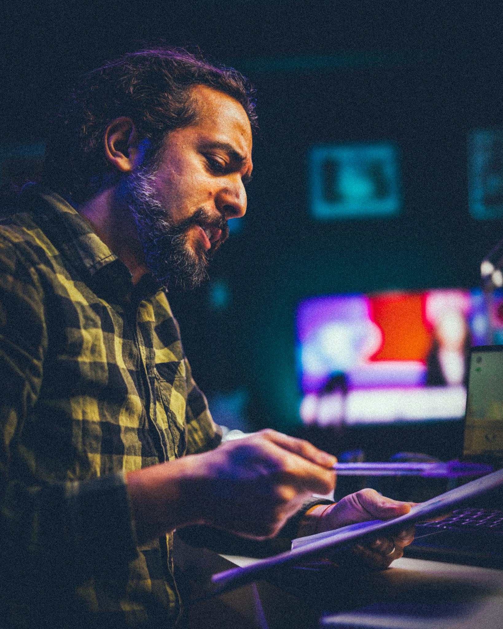 Man in a green checkered shirt reviewing papers with a pen in a dimly lit room with a blurred screen in the background.