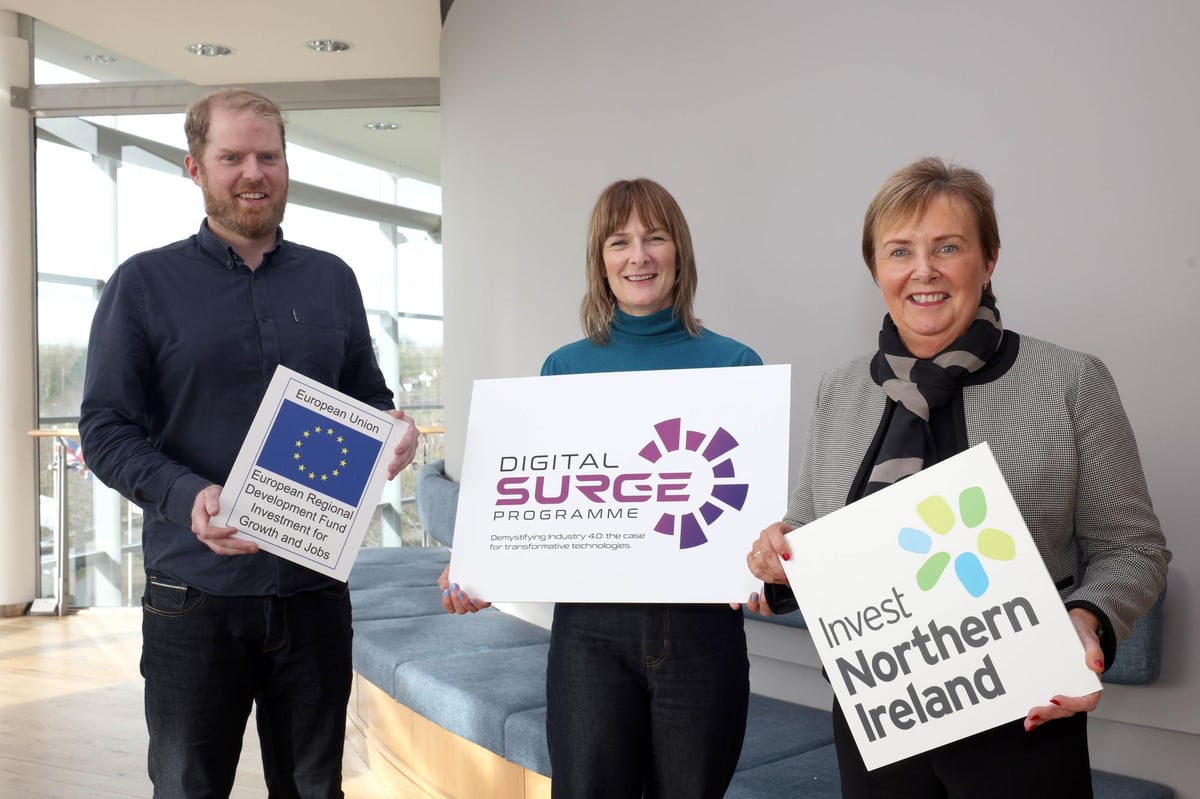 Three people indoors holding signs for European Union, Digital Surge Programme, and Invest Northern Ireland.