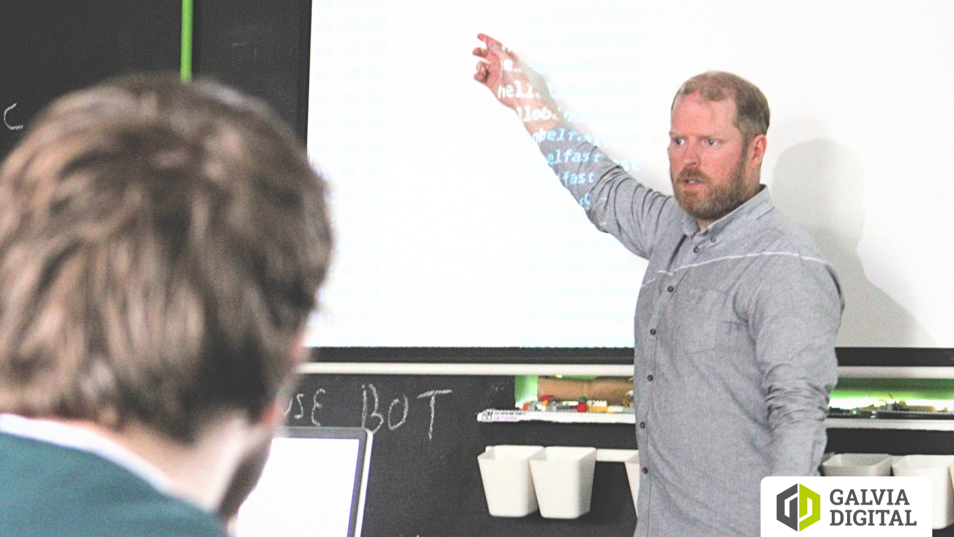 Man with beard pointing at a projection screen with code during a presentation in a classroom setting.