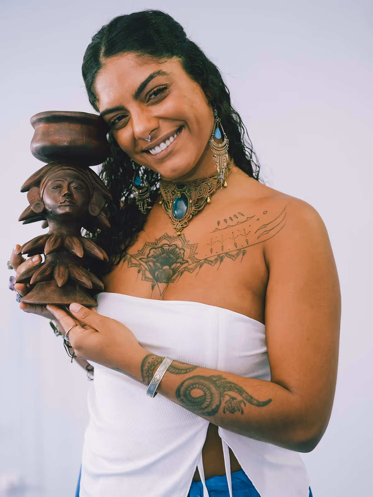 Mulher sorridente com tatuagens e joias segurando uma escultura de madeira com forma feminina.