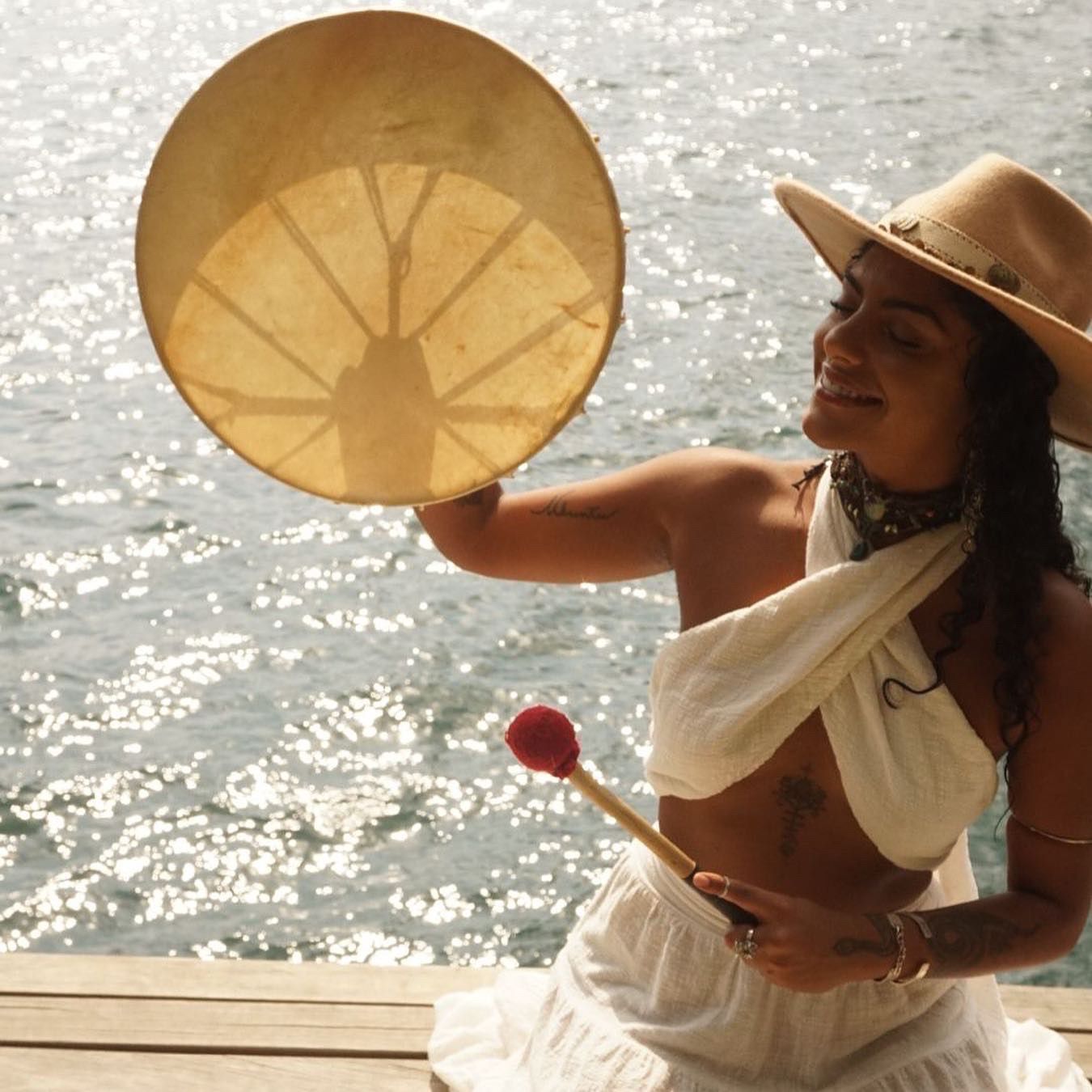 Mulher sorrindo tocando um tambor circular com uma baqueta vermelha, sentada em um deck de madeira à beira da água.