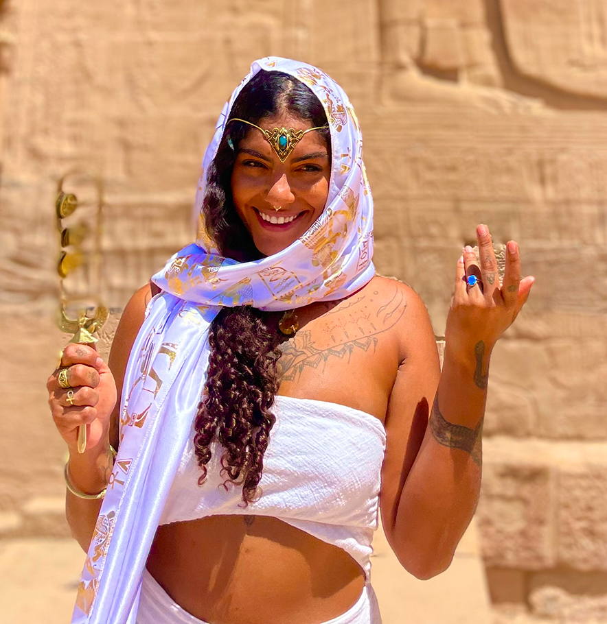 Mulher sorridente com véu branco decorado com símbolos dourados, vestindo ornamentos e tatuagens, em frente a uma parede de pedra.