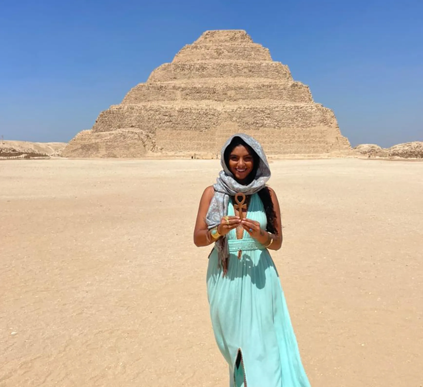 Mulher sorridente de vestido azul claro e lenço na cabeça segurando um símbolo antigo na frente de uma pirâmide egípcia de degraus sob céu azul claro.